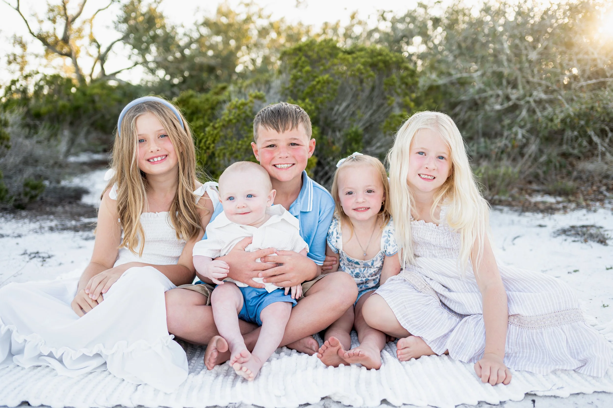 Group of five children, four girls and one boy, sitting on a beach blanket with greenery and bushes in the background during sunset.