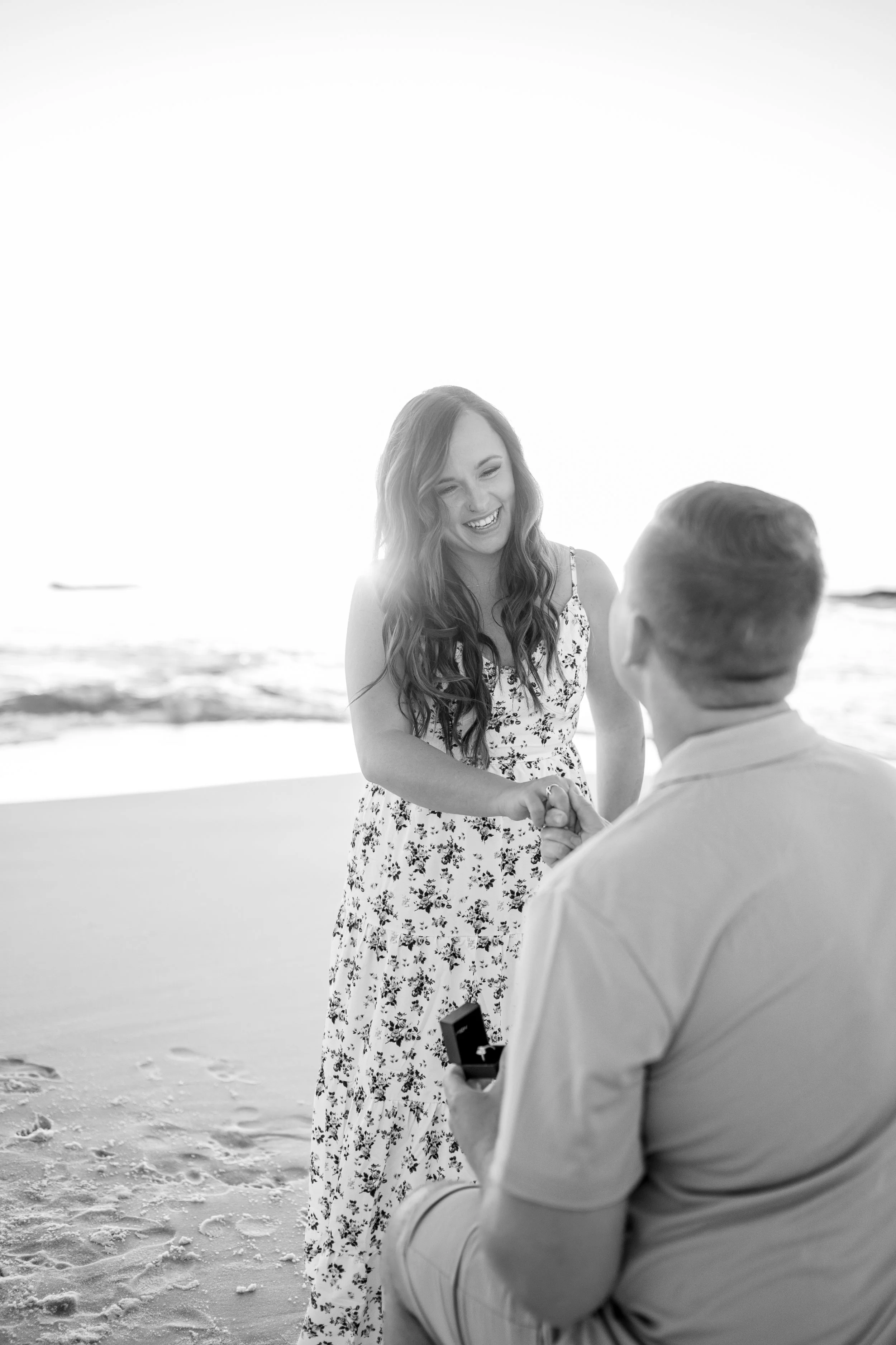 A woman in a floral dress is smiling and holding hands with a man at the beach, who is kneeling and holding a box, likely during a proposal.