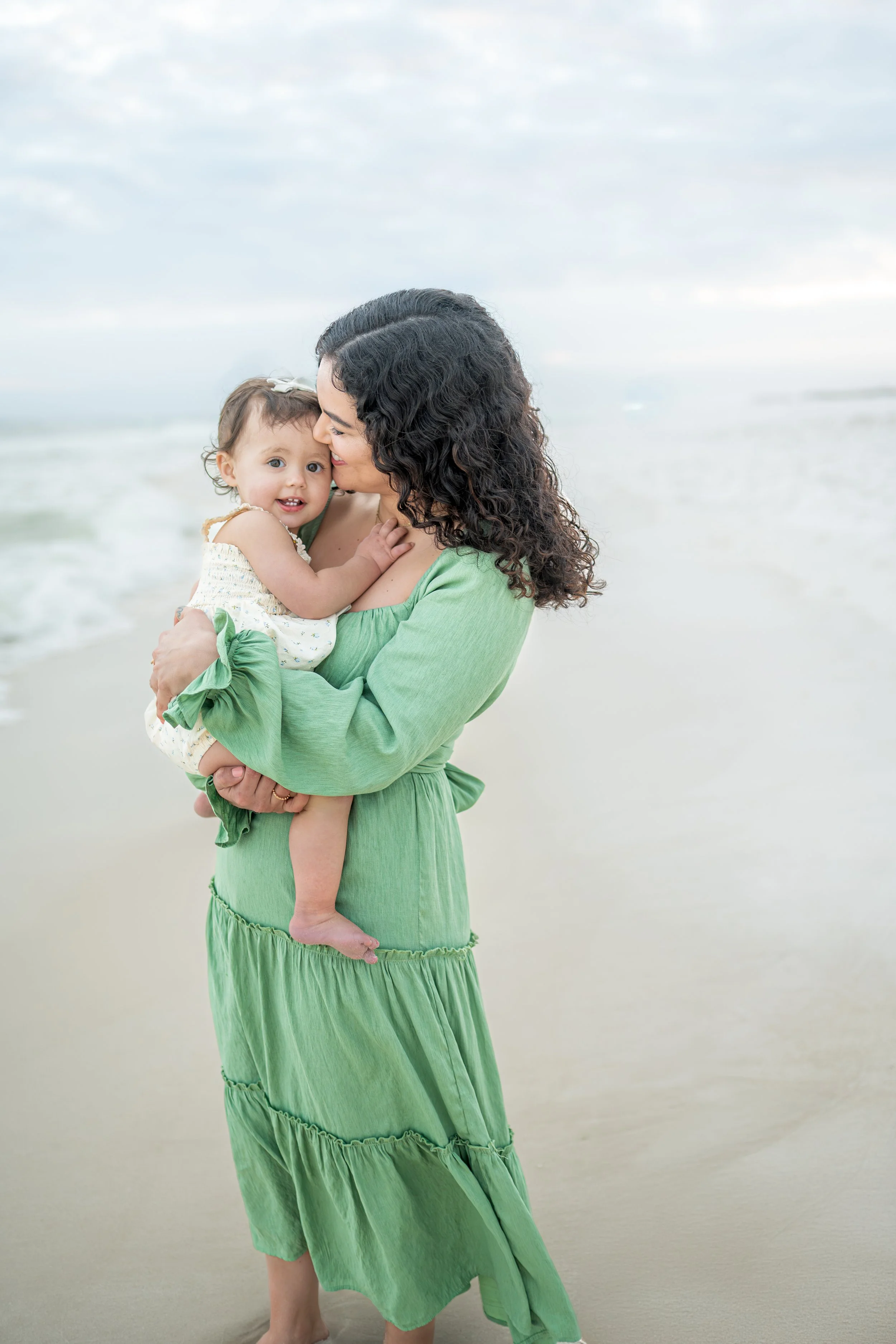 A woman in a green dress holding a young girl in white on a beach.