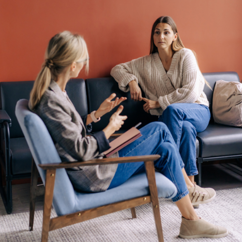 A woman in a gray blazer and blue jeans talks to another woman in a beige sweater and blue jeans in a waiting room.