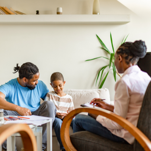 A family sitting together in a living room, with a man, a woman, and a boy. The man and boy are smiling, and the woman is taking notes or filling out a form.
