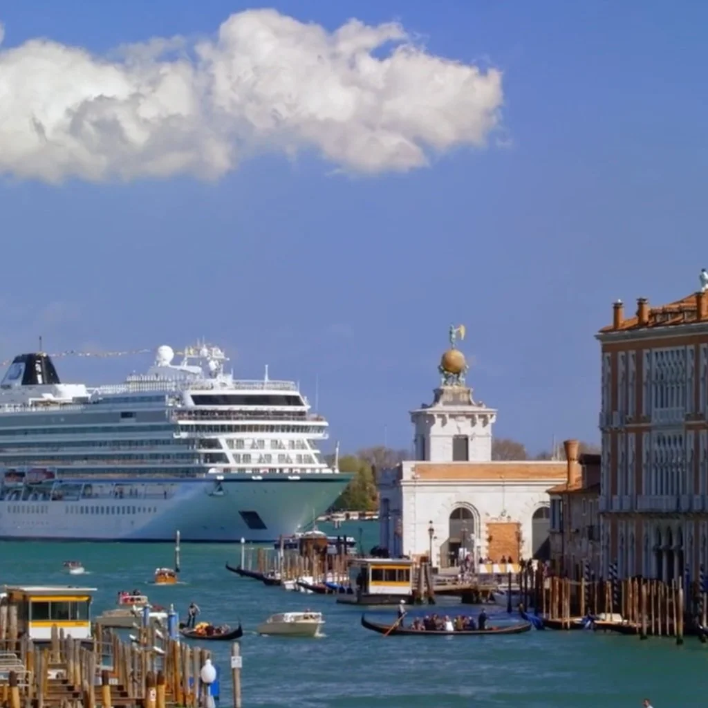 View of a cruise ship docked in Venice's waterfront near historic buildings and gondolas on the water under a partly cloudy sky.