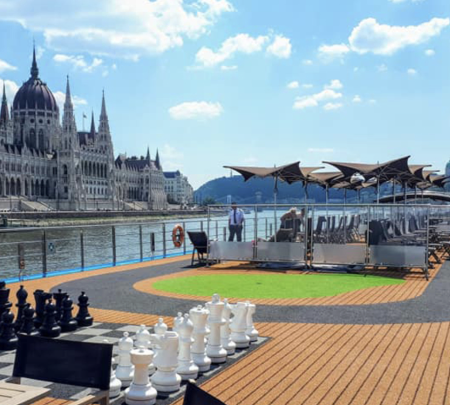 View of a river with a historic castle-style building on the left, a terrace with umbrellas and seating in the foreground, and a giant chess set on the street, under a partly cloudy sky.