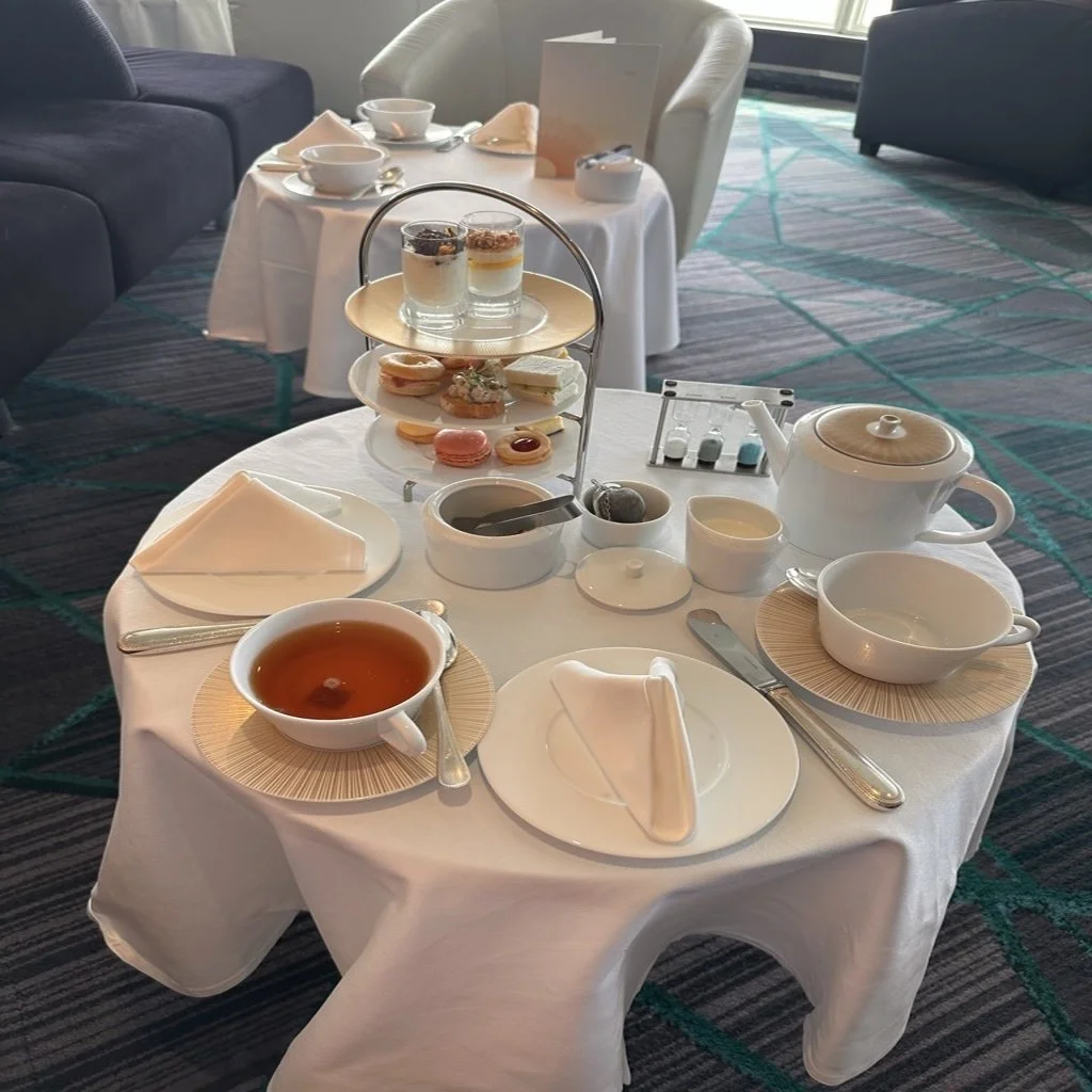 A table set for afternoon tea with a white tablecloth, a teapot, a cup of tea, two bowls, and a three-tier stand holding assorted finger sandwiches, pastries, and desserts. In the background, there are additional tables and chairs in a well-lit room.