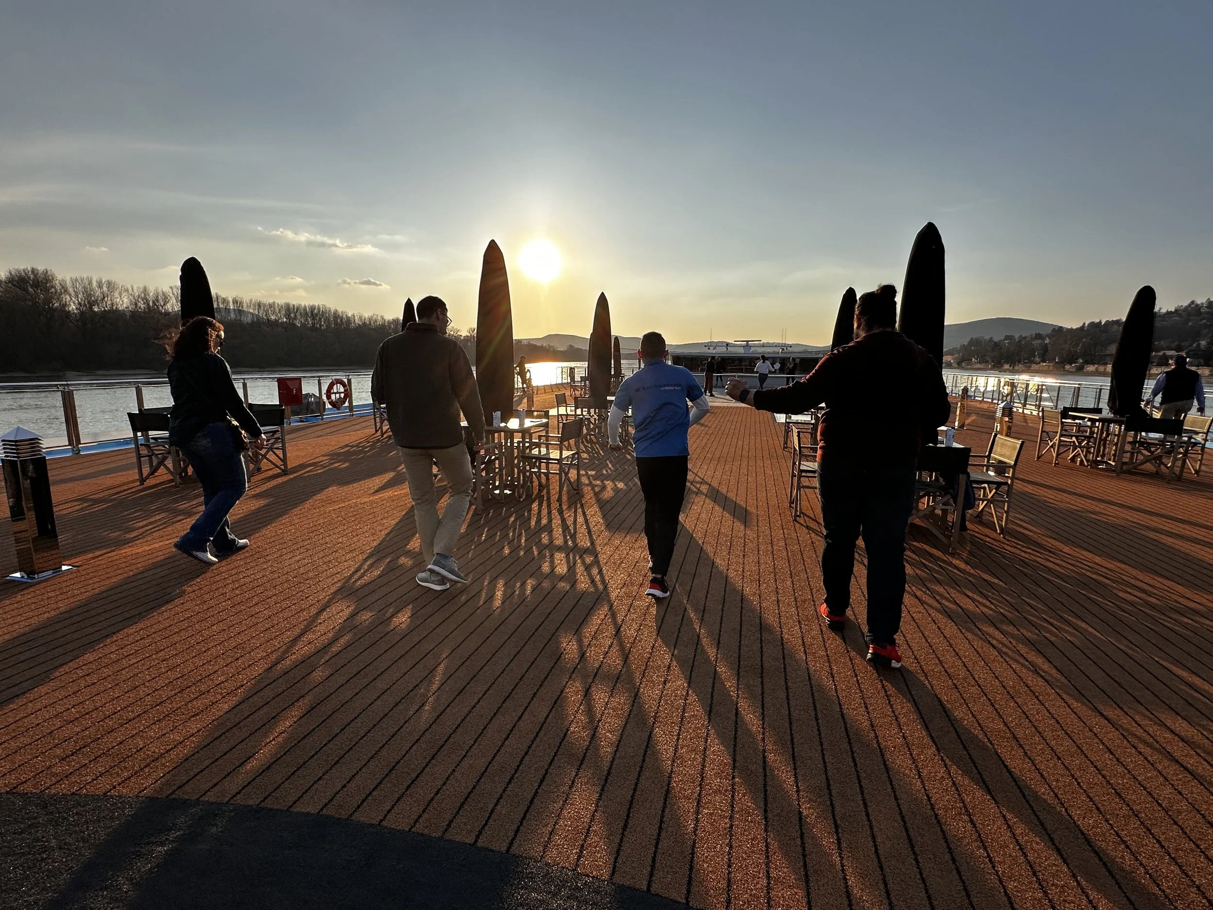 People walking on the deck of a boat during sunset with water and hills in the background.