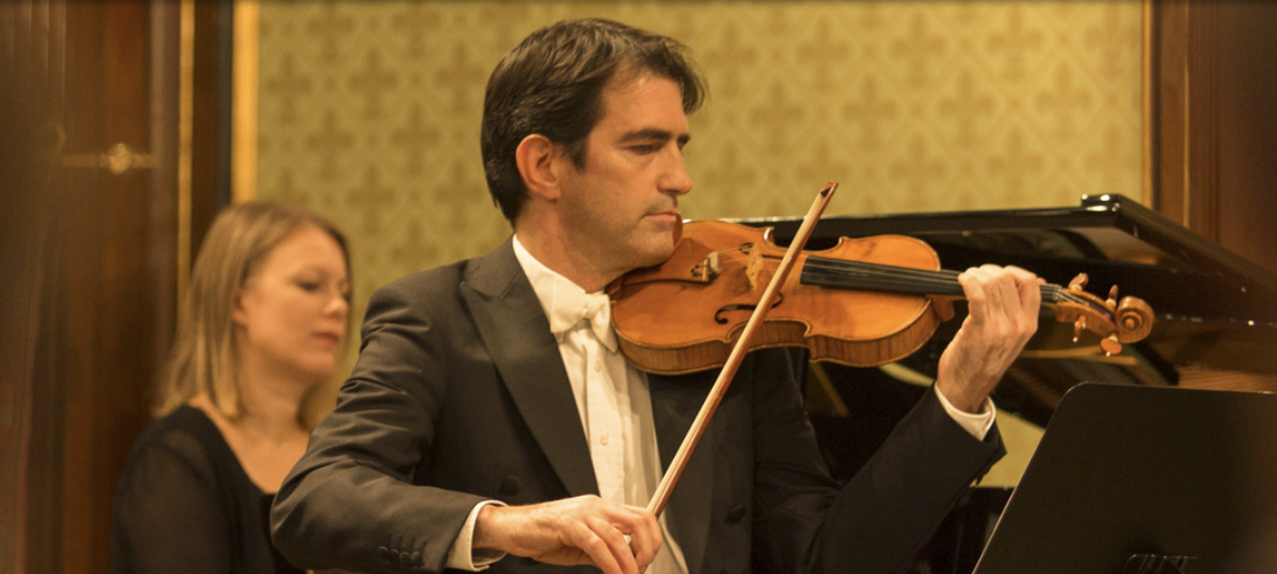A man in formal attire playing the violin in a concert hall, with a woman in the background.