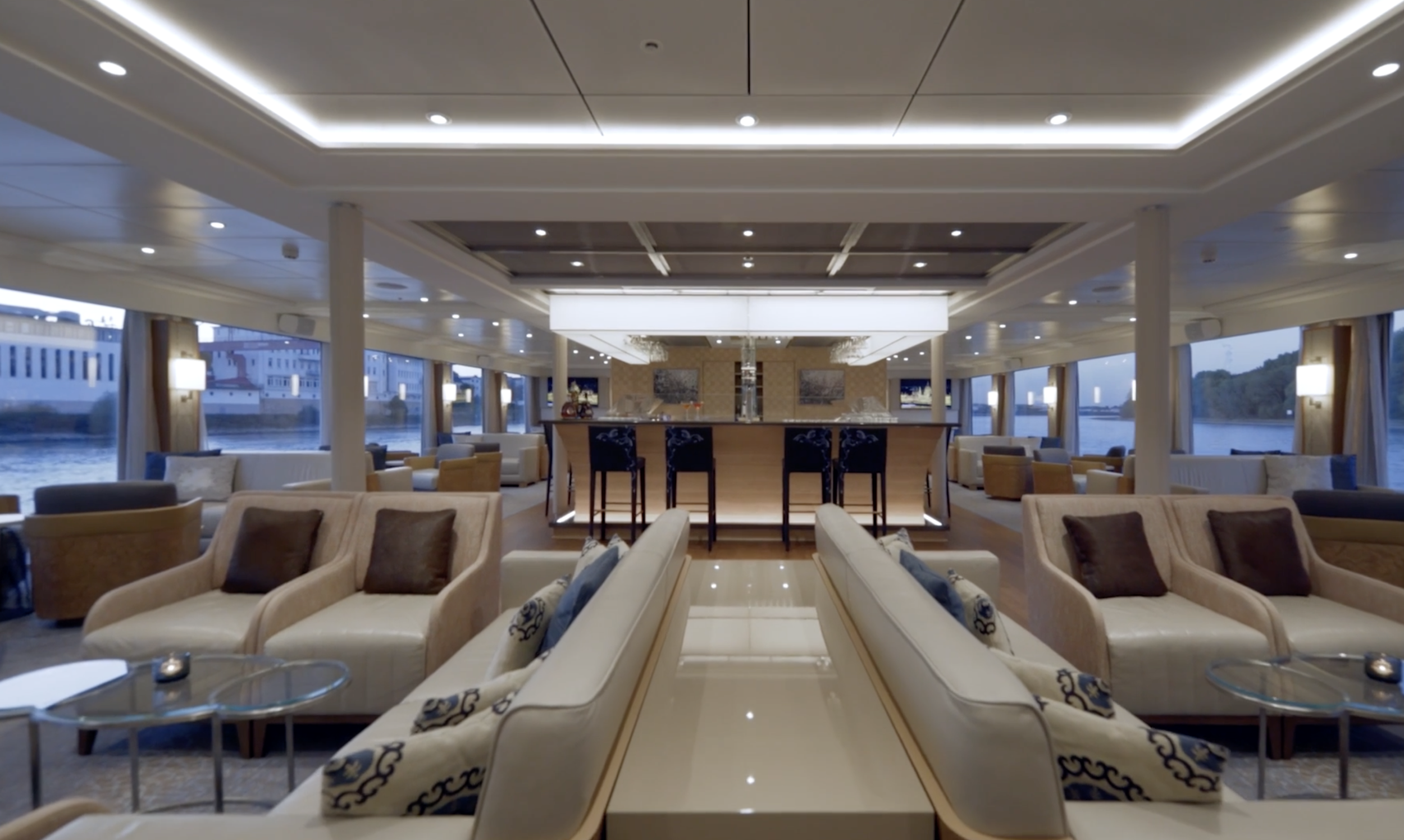 Interior of a modern lounge area with white sofas, armchairs, and round glass tables, overlooking a waterfront view through large windows, with a bar in the center.