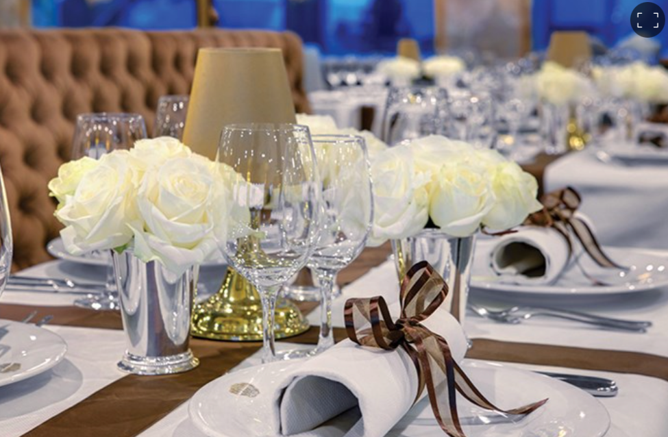 Elegant table setting with white roses in silver vases, white napkins with brown ribbons, wine glasses, and a gold lamp on a brown tablecloth.