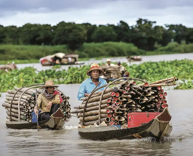 Two small boats filled with bamboo poles, carrying two women dressed in typical clothing, floating on a river with green vegetation and more boats in the background.