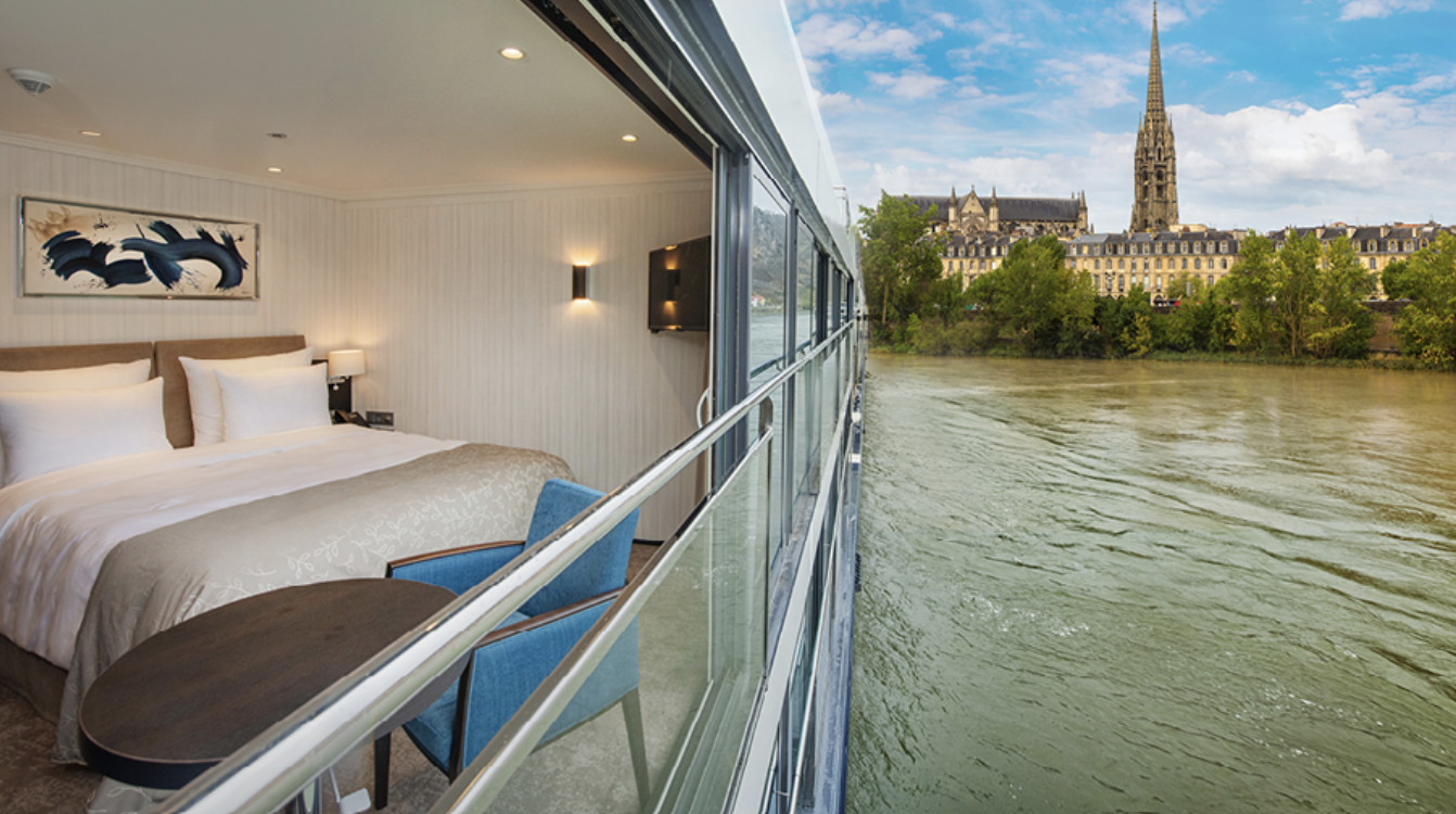 Interior of a hotel room with a bed, artwork, and a small table next to a large window. The window opens to a view of a river and a city with a church steeple and historic buildings.