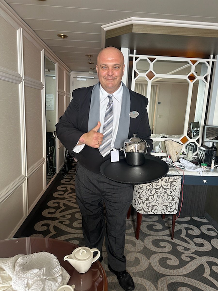 A man in a suit holding a tray with a silver teapot in a hotel room, smiling and giving a thumbs-up.