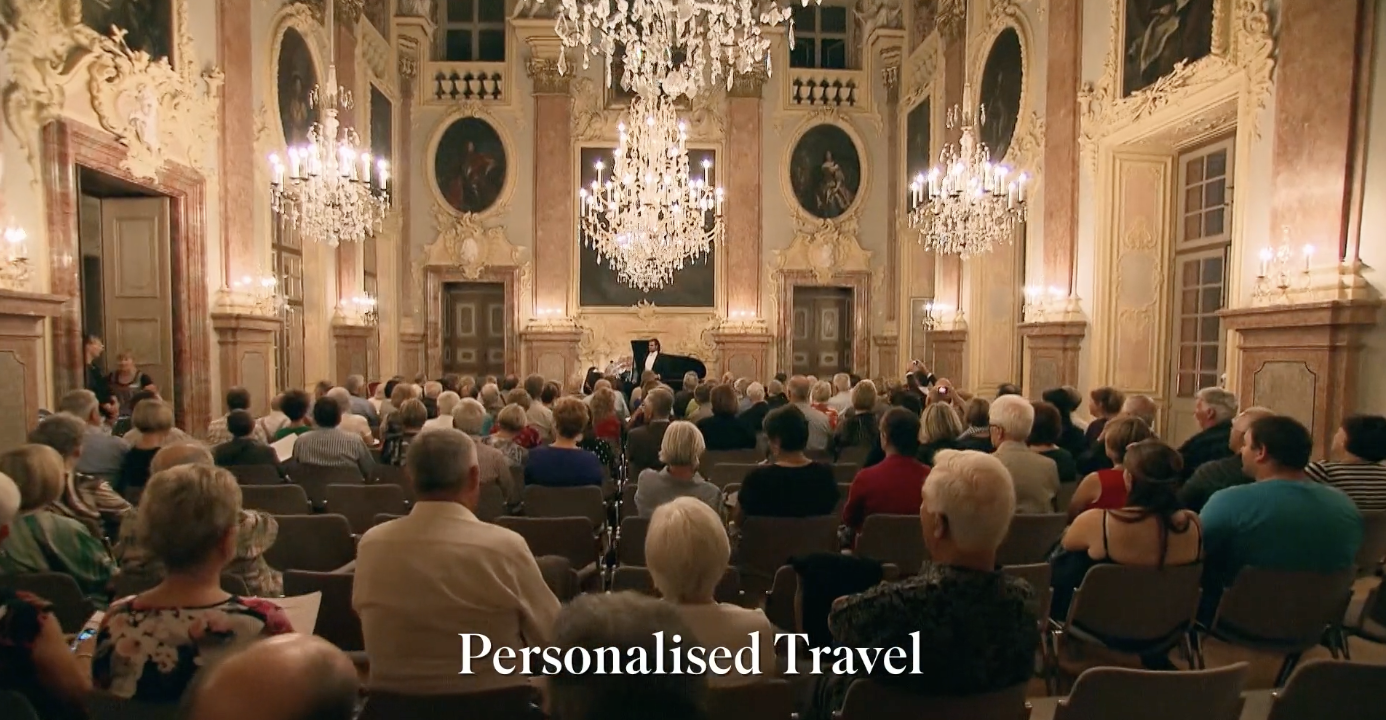 An audience seated in a luxurious, ornate ballroom with gold accents and chandeliers, watching a pianist perform at the front.