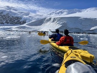 Two people kayaking in an icy, glacier-filled landscape with snow-covered mountains in the background.