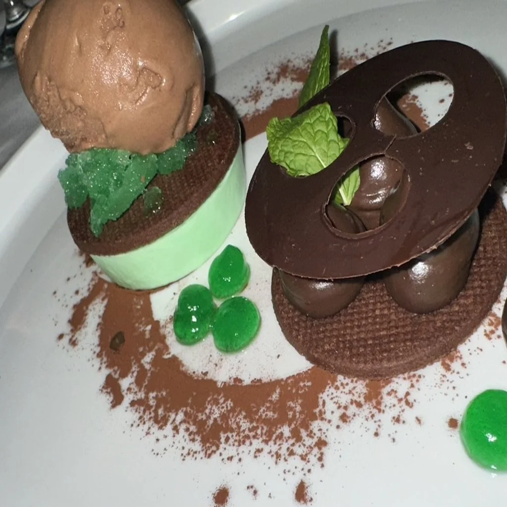 Decorative chocolate dessert with green jelly beans, mint leaf, and cocoa powder on a white plate.