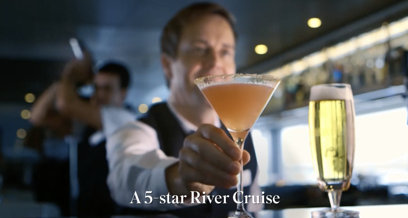 A person holding a pink cocktail in a glass, with a glass of beer beside it, on a boat or cruise ship, with the text "A 5-star River Cruise" at the bottom.
