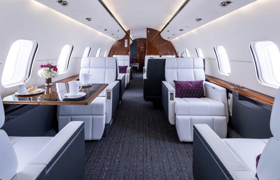 Interior of a luxurious airplane cabin with white leather seats, wooden tables set for breakfast, and windows along both sides.