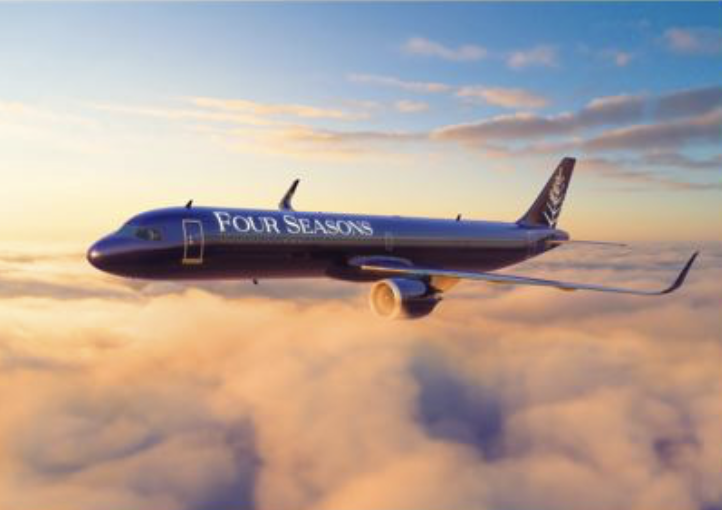 A Boeing airplane with the Four Seasons logo flying above the clouds at sunset.