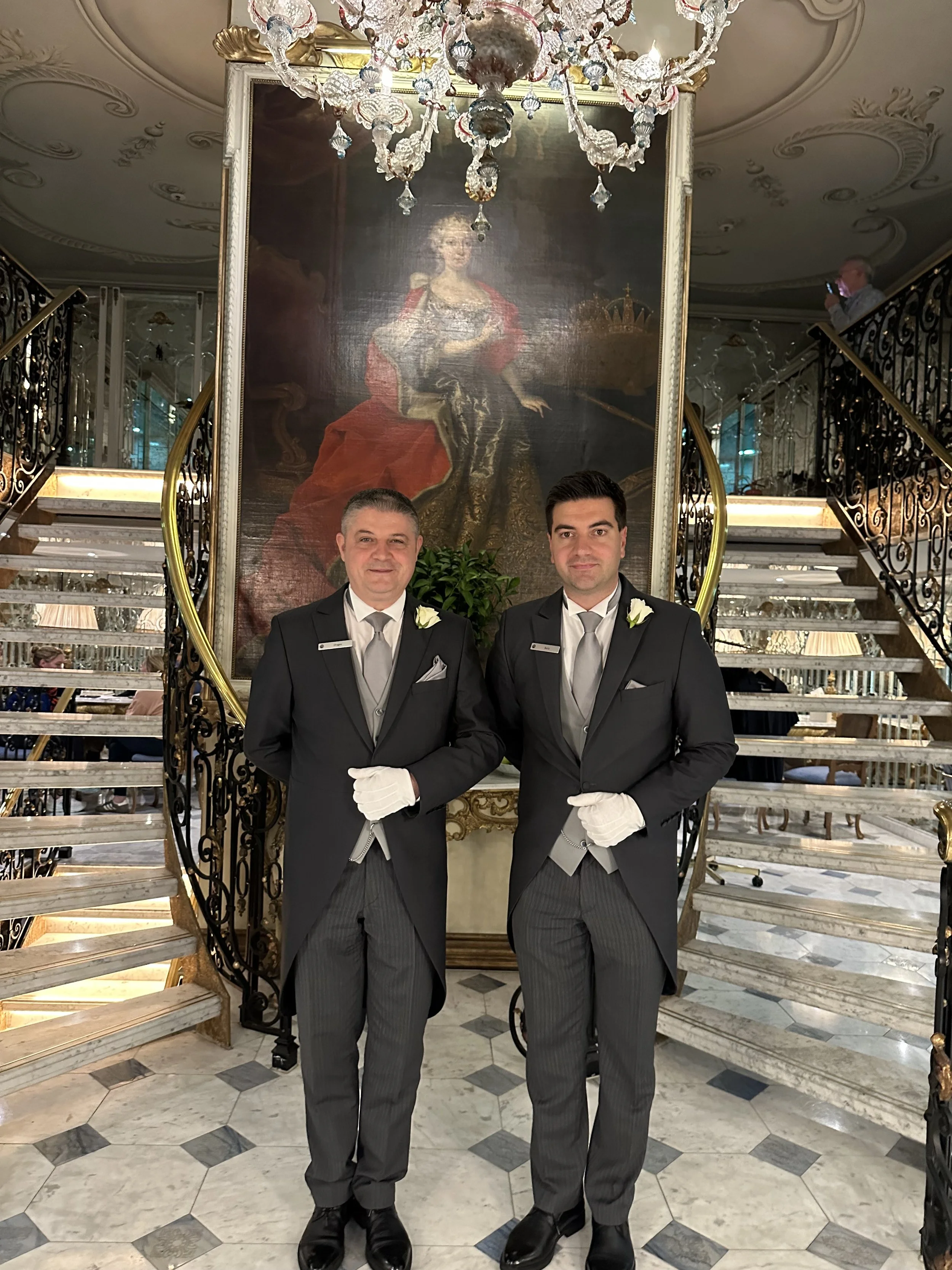 Two men dressed in black tuxedos and white gloves standing in front of a large portrait of a historical woman in a red and gold dress, inside an ornate, elegant building with a chandelier and marble staircase.