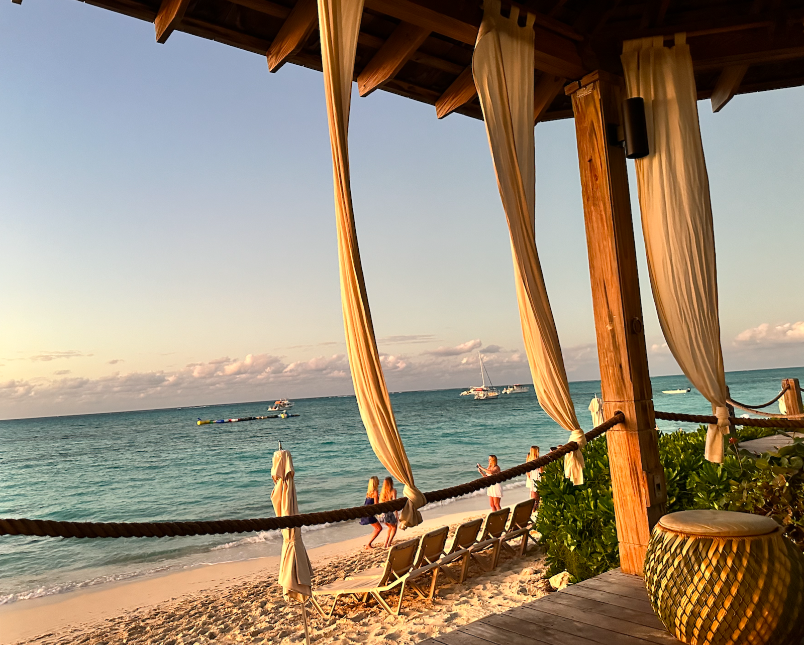 A beach scene viewed from a shaded wooden cabana with beige curtains. There are lounge chairs on the sand, a few people walking and taking photos, with the ocean and boats in the background. The sky is clear with some clouds, and the scene is bathed in warm sunlight.