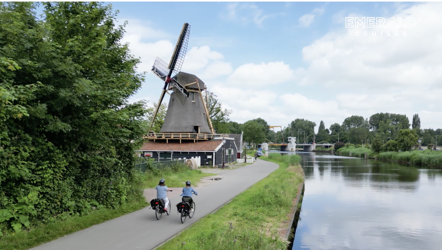 Two people cycling along a river, with a windmill and a bridge in the background.