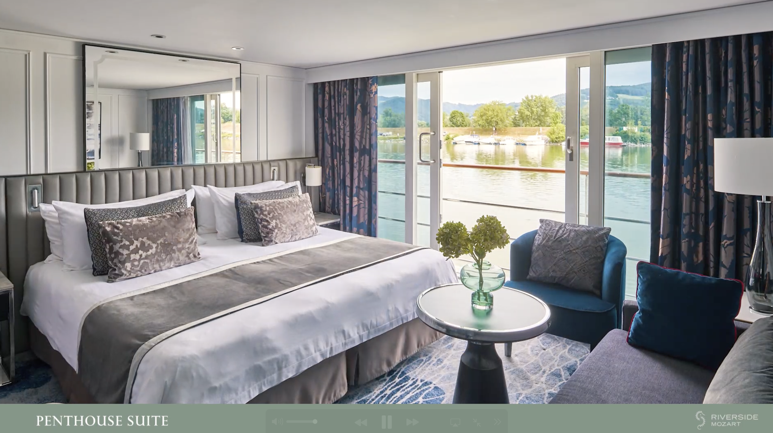 A bedroom with a large bed, decorative pillows, a mirror, and view of a river with boats outside the window.
