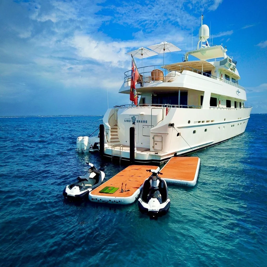 Large white yacht with a wooden dock and two jet skis attached, on calm ocean water under a partly cloudy sky.