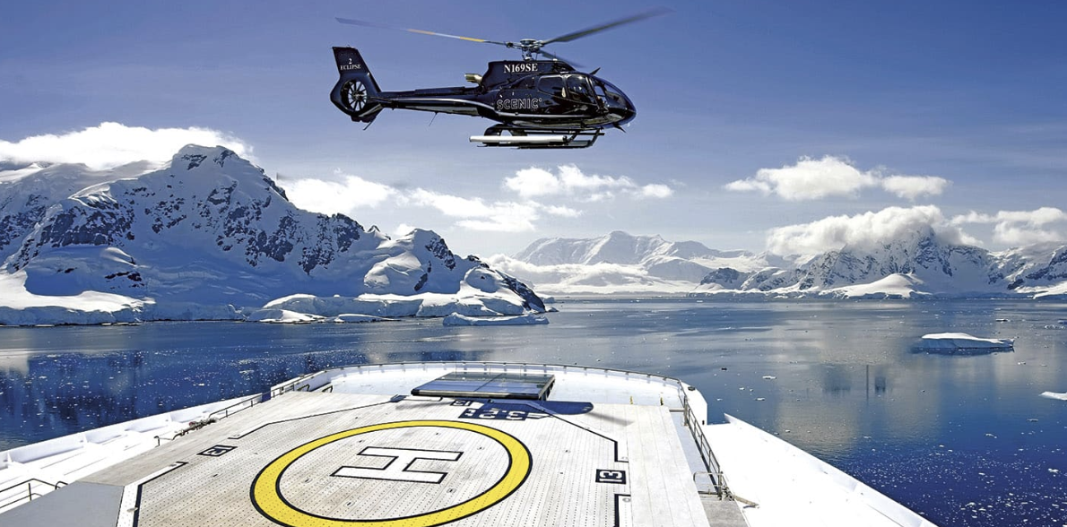 A helicopter flying over a helipad on a ship in icy waters with snow-covered mountains in the background.