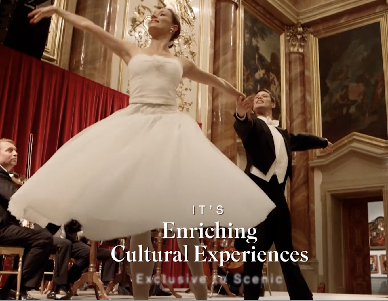A woman in a white dress and a man in a tuxedo dancing on stage during a cultural performance, with a string quartet and ornate decor in the background.