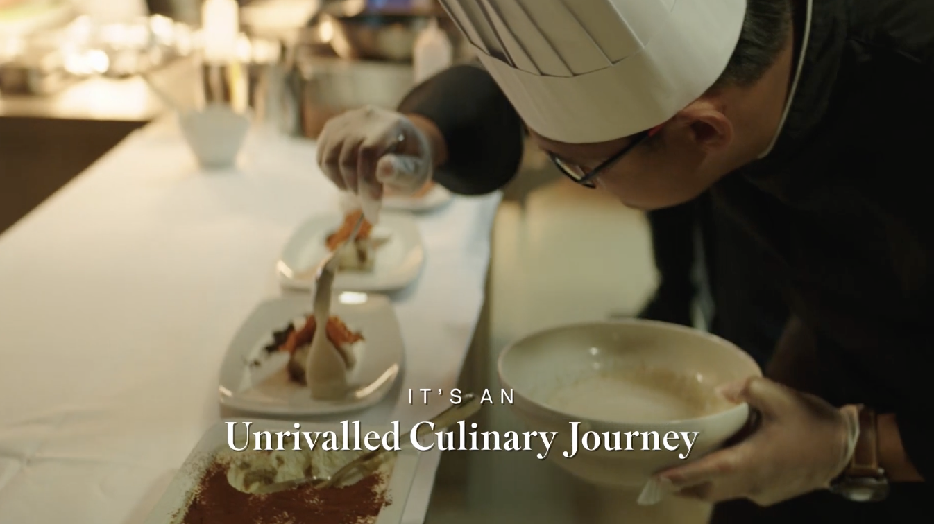 A chef in a white hat and black uniform is carefully plating a dish in a professional kitchen. Several plated dishes are lined up on the counter, with one being finished by the chef. The scene highlights a culinary process.