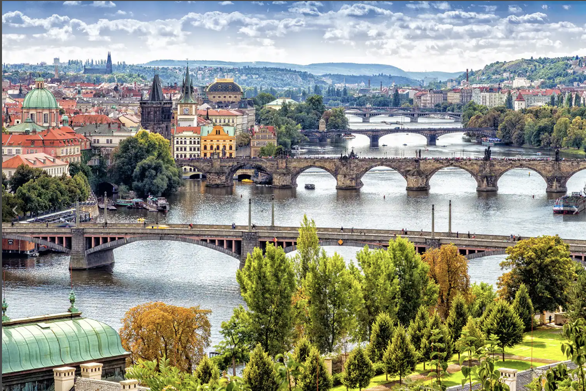 A panoramic view of Prague with multiple bridges crossing the Vltava River, historic buildings, and green trees in the foreground and background.