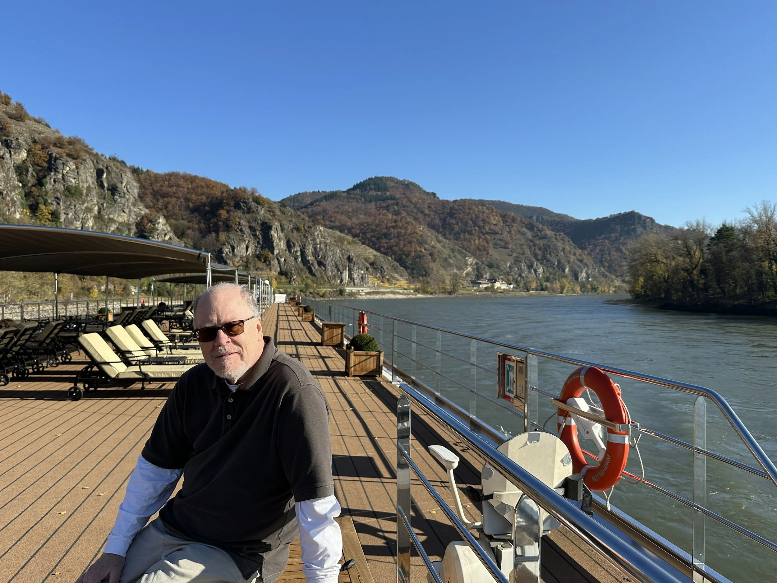 A man wearing sunglasses and a black long sleeve shirt with white sleeves sitting on a deck beside a river with mountains and trees in the background on a sunny day.