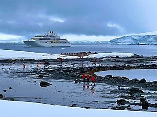 A large cruise ship docked near a snow-covered shoreline with ice and rocks, under overcast skies.