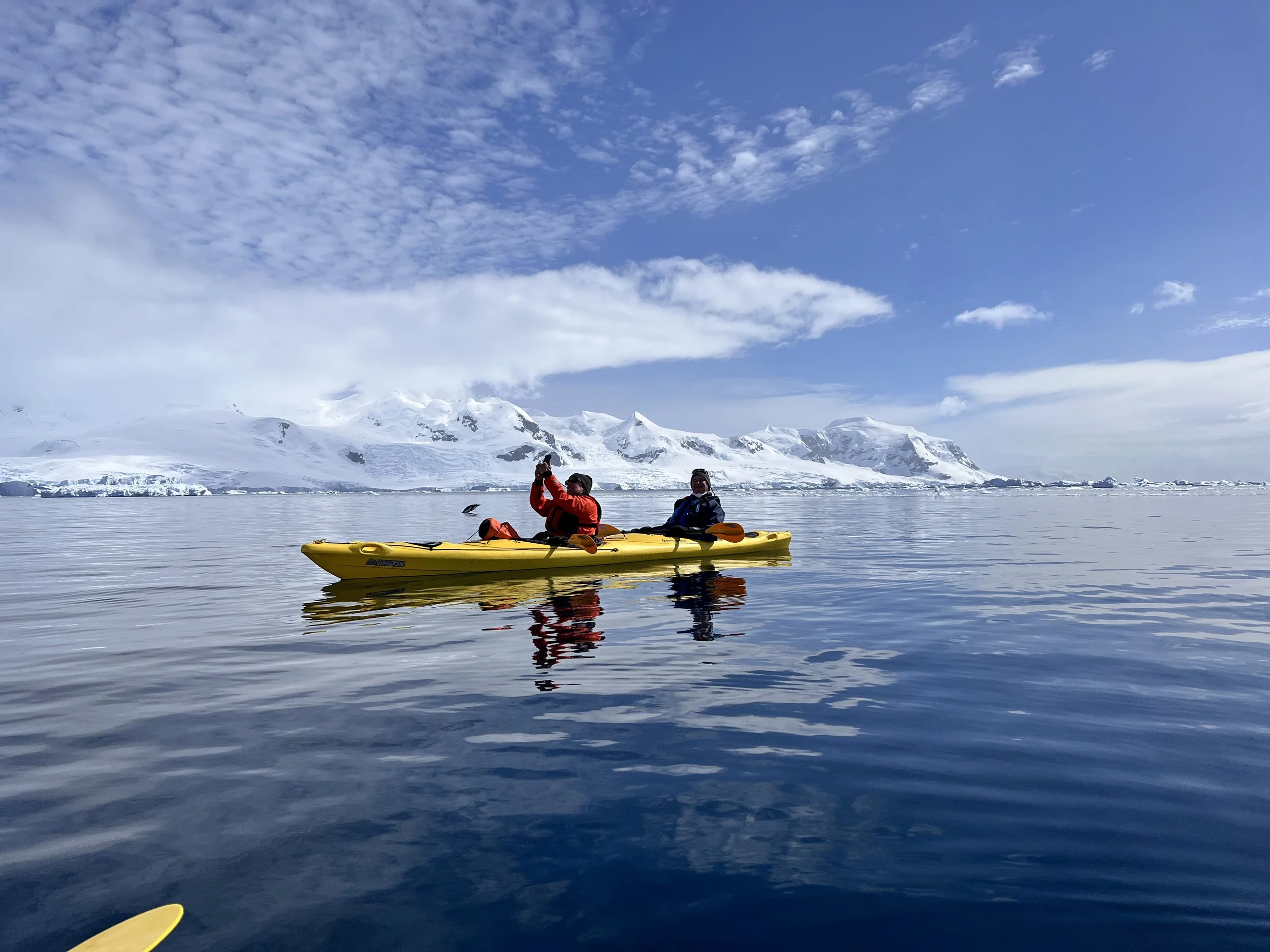 Two people in a yellow kayak on calm icy water with snowy mountains and blue sky in the background.