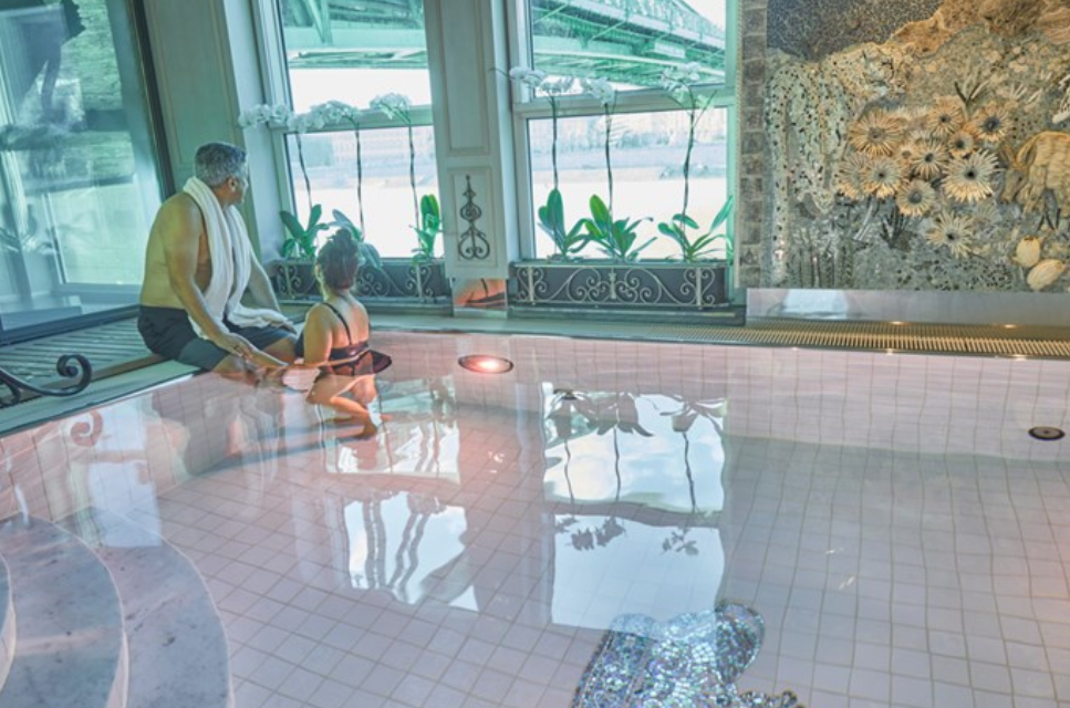 An adult and a child sitting by the edge of an indoor swimming pool, with large windows and decorative wall art in the background.