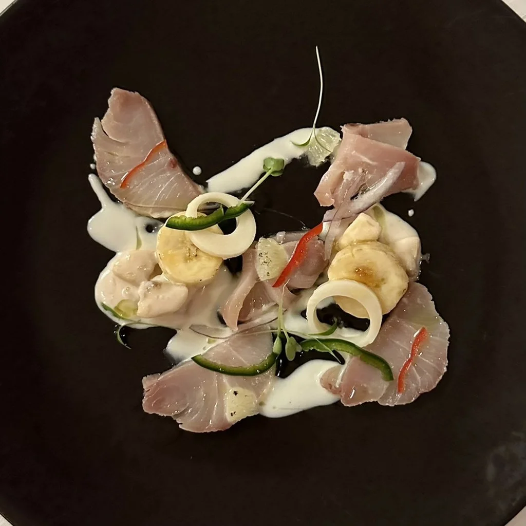 A plate of raw fish sashimi with slices of fish, banana, green chili, red chili, onion, and microgreens, served on a black plate with white sauce.