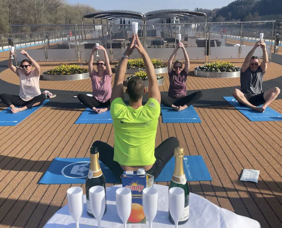 A group of four women and one man participating in a yoga class on a boat deck, sitting on yoga mats and holding smartphones above their heads. A man, facing away from the camera, sits in front of them, leading the session. There are flowers, champagne bottles, glasses, and snacks on a table in the foreground. The weather is sunny with clear skies.