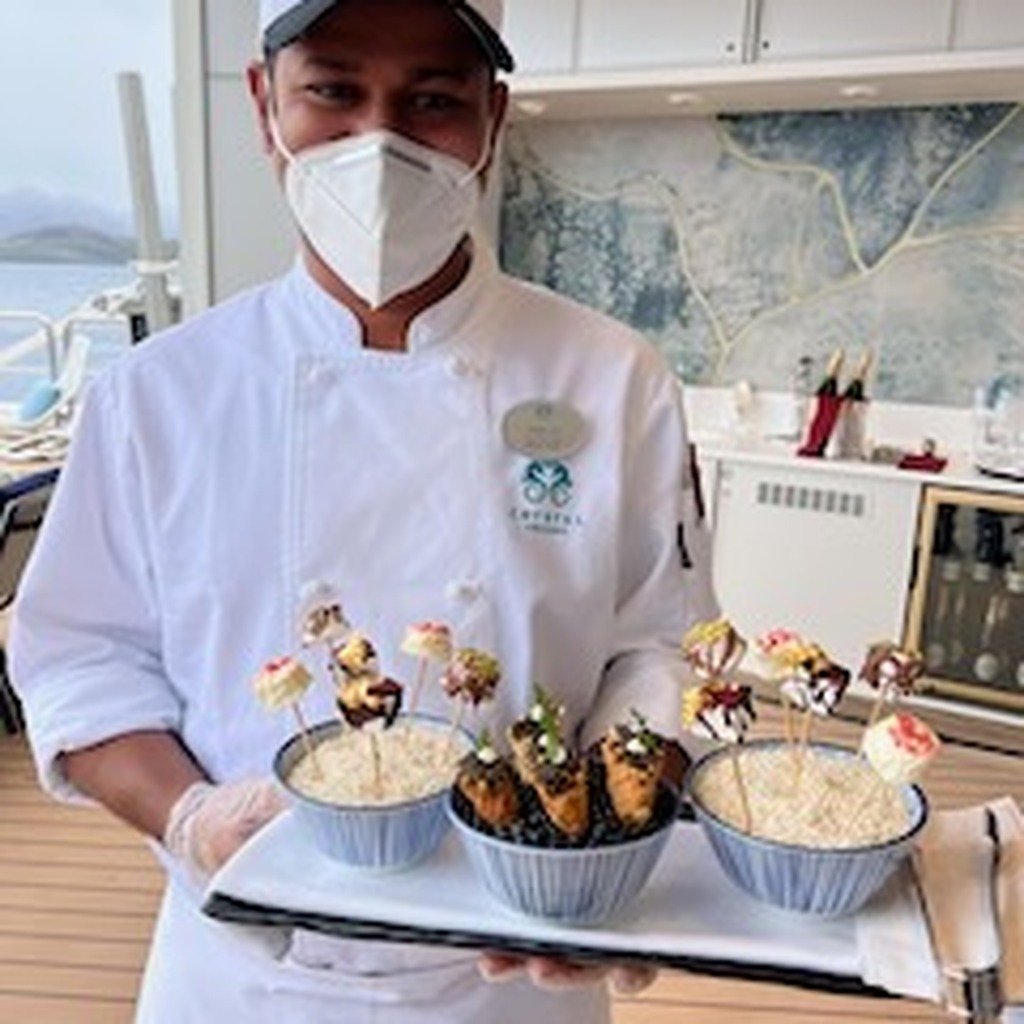 Chef in a white uniform and face mask holding a tray with bowls of decorated desserts.