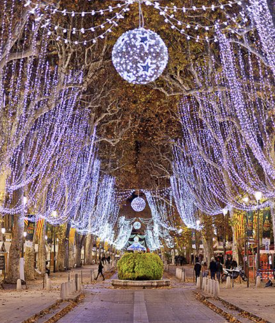 Street decorated with purple and white Christmas lights and hanging ornaments, with trees and people walking.
