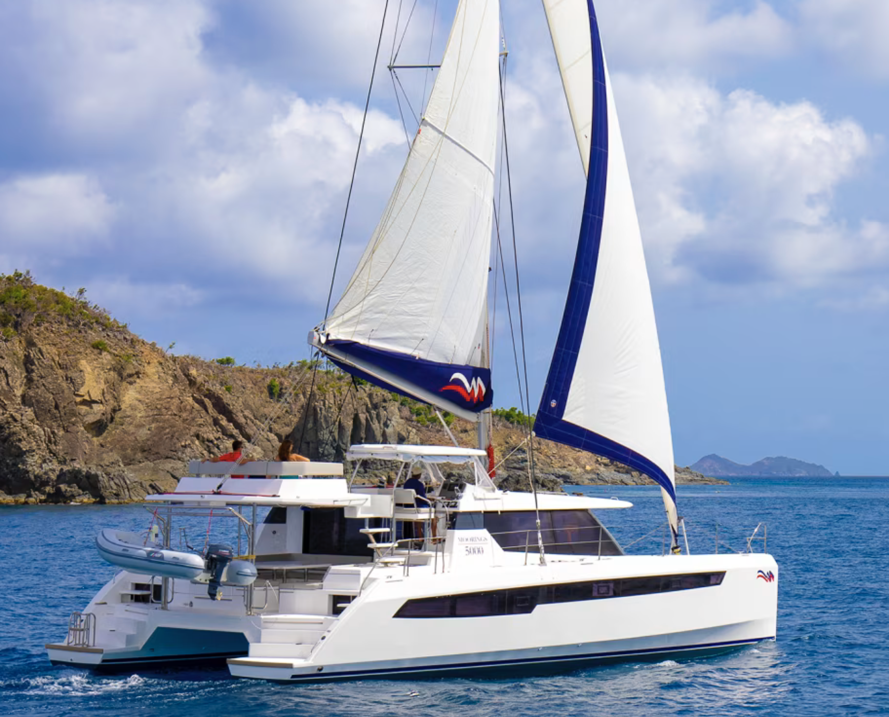 A white sailing yacht with blue sails on the ocean near rocky islands under a partly cloudy sky.