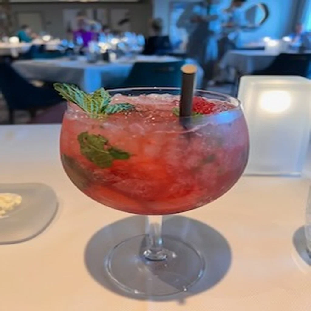 A large glass of red cocktail garnished with mint leaves and fruit, on a white table in a restaurant setting.
