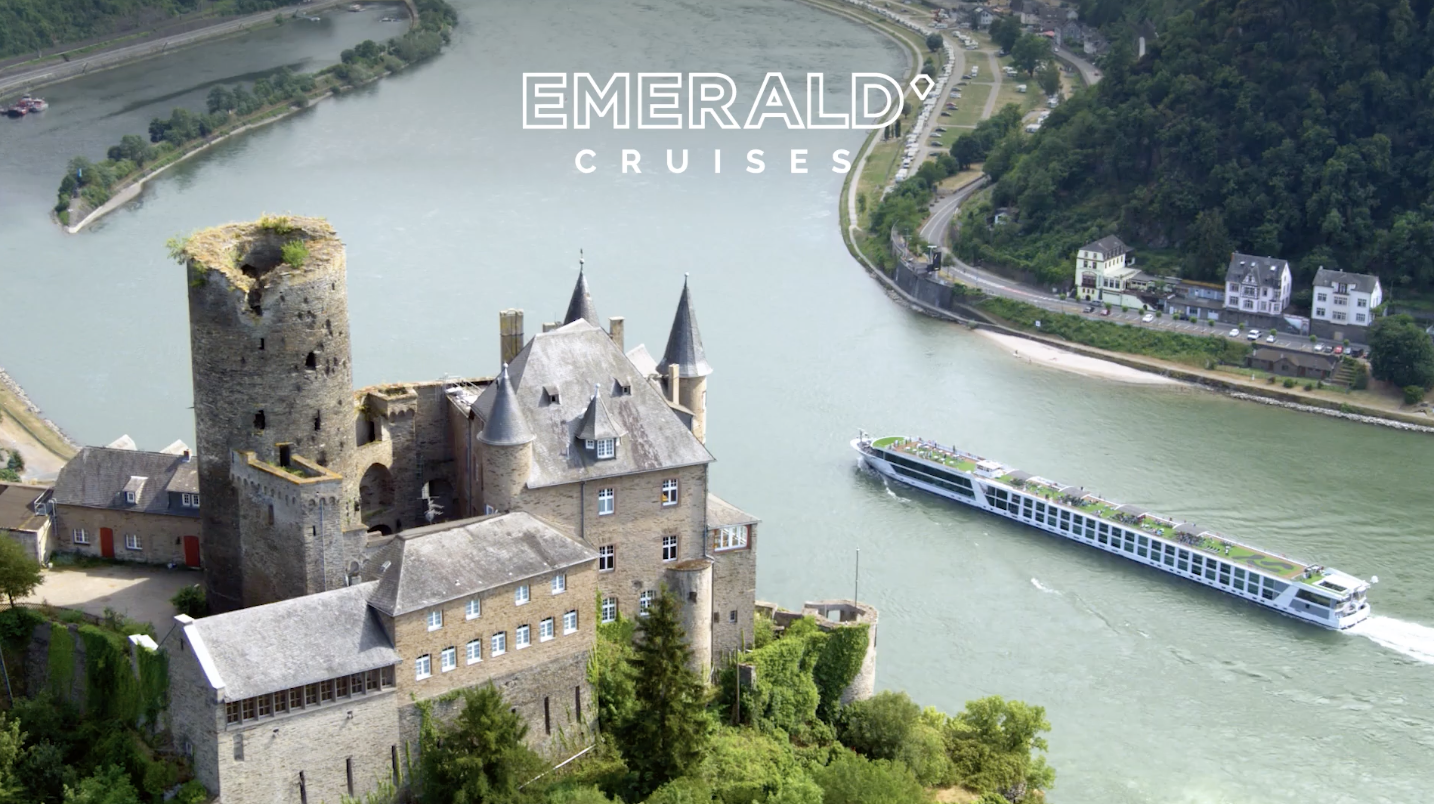 Aerial view of a castle on a river with a cruise boat passing by and green hills surrounding the area. The image includes the emerald cruises logo at the top.
