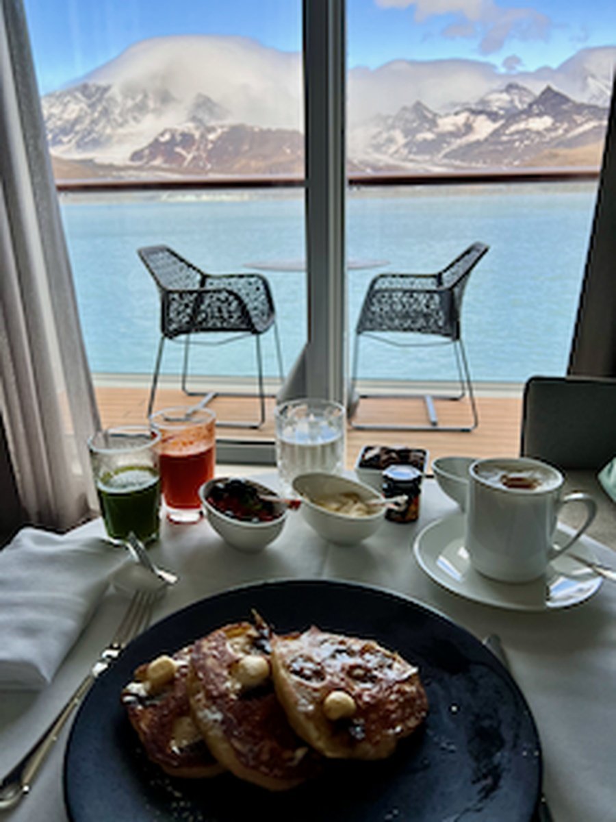 A breakfast table with pancakes topped with butter, and various drinks including green, red, and transparent liquids, set near a large window overlooking a lake with mountains in the background.