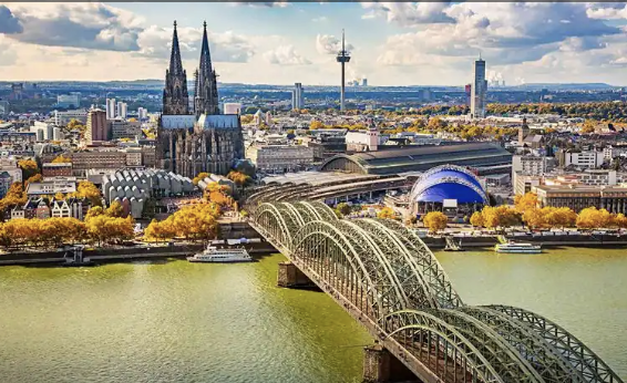 A cityscape of Cologne, Germany, featuring the Cologne Cathedral, the Hohenzollern Bridge, and the Rhine River with boats.