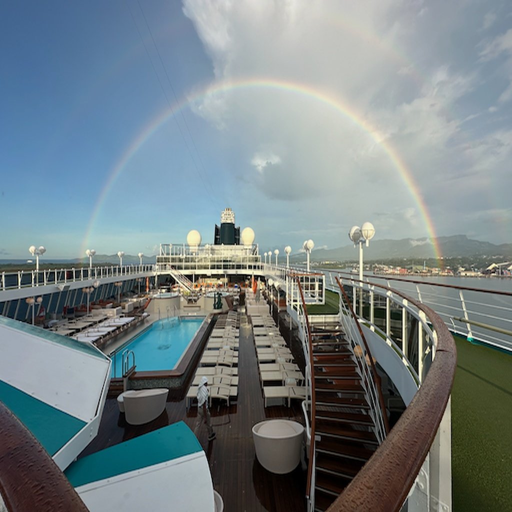 View of the top deck of a cruise ship with a rainbow in the sky, a swimming pool, lounge chairs, and a few people, with a cloudy sky and distant land in the background.
