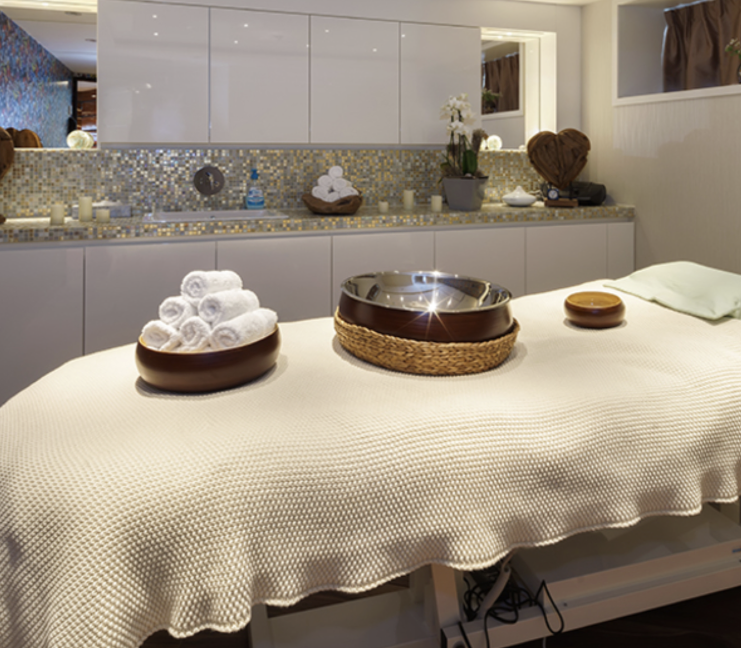 Spa massage table covered with a textured beige cloth, with rolled white towels, a brown bowl, and a small wooden dish on top. In the background, a bathroom area with a mosaic tile backsplash, bottles, and decorative items.