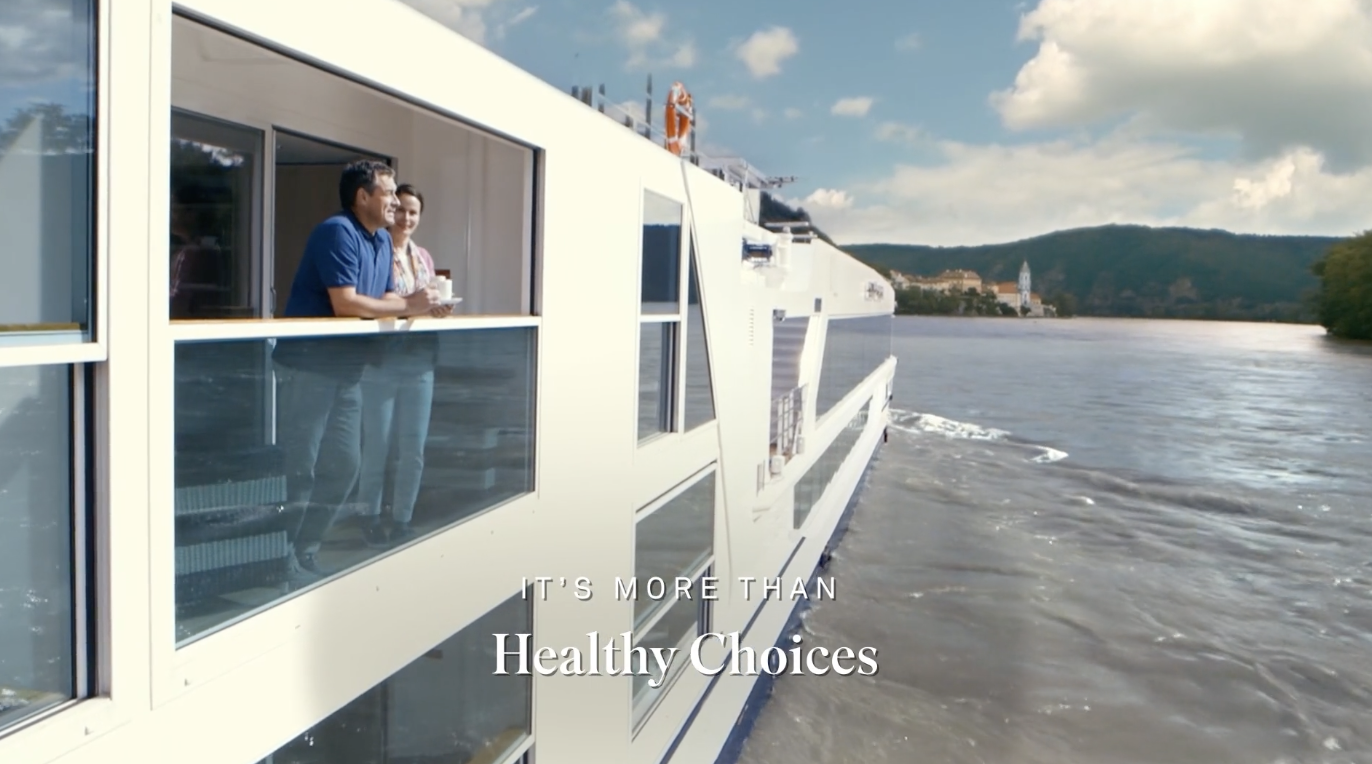 Two people standing on the deck of a white boat or cruise ship, looking out over the water, with a scenic landscape of hills and a small town in the background.