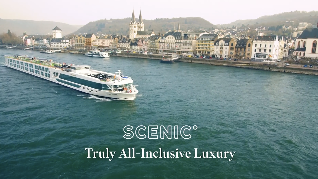 A large white and green cruise ship sailing on a river with a European town and hills in the background. The text on the image reads 'SCENIC Truly All-Inclusive Luxury.'