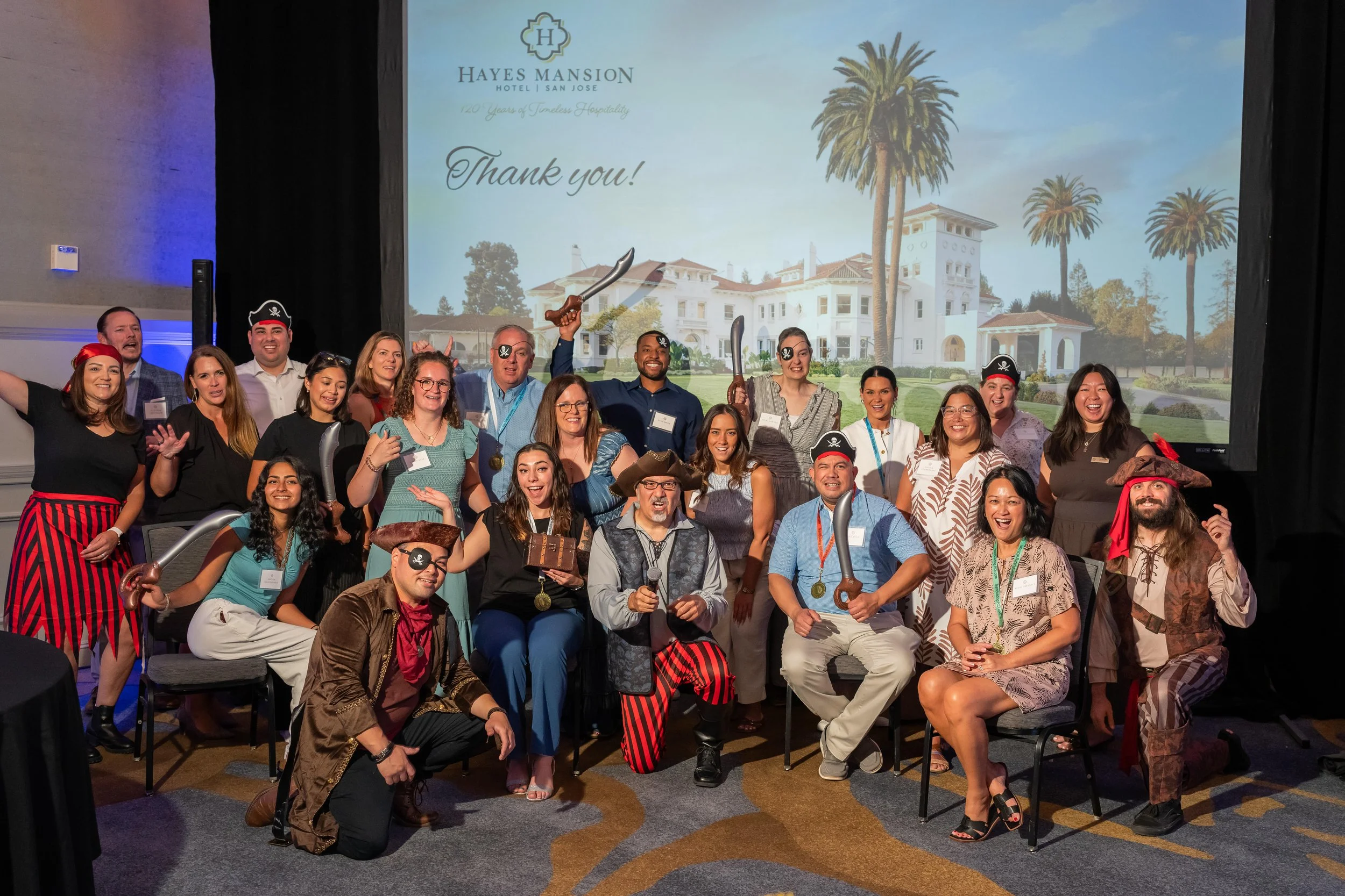 Group of people posing with pirate accessories such as eye patches and swords at a celebratory event in front of a large screen displaying a hotel and thank you message.