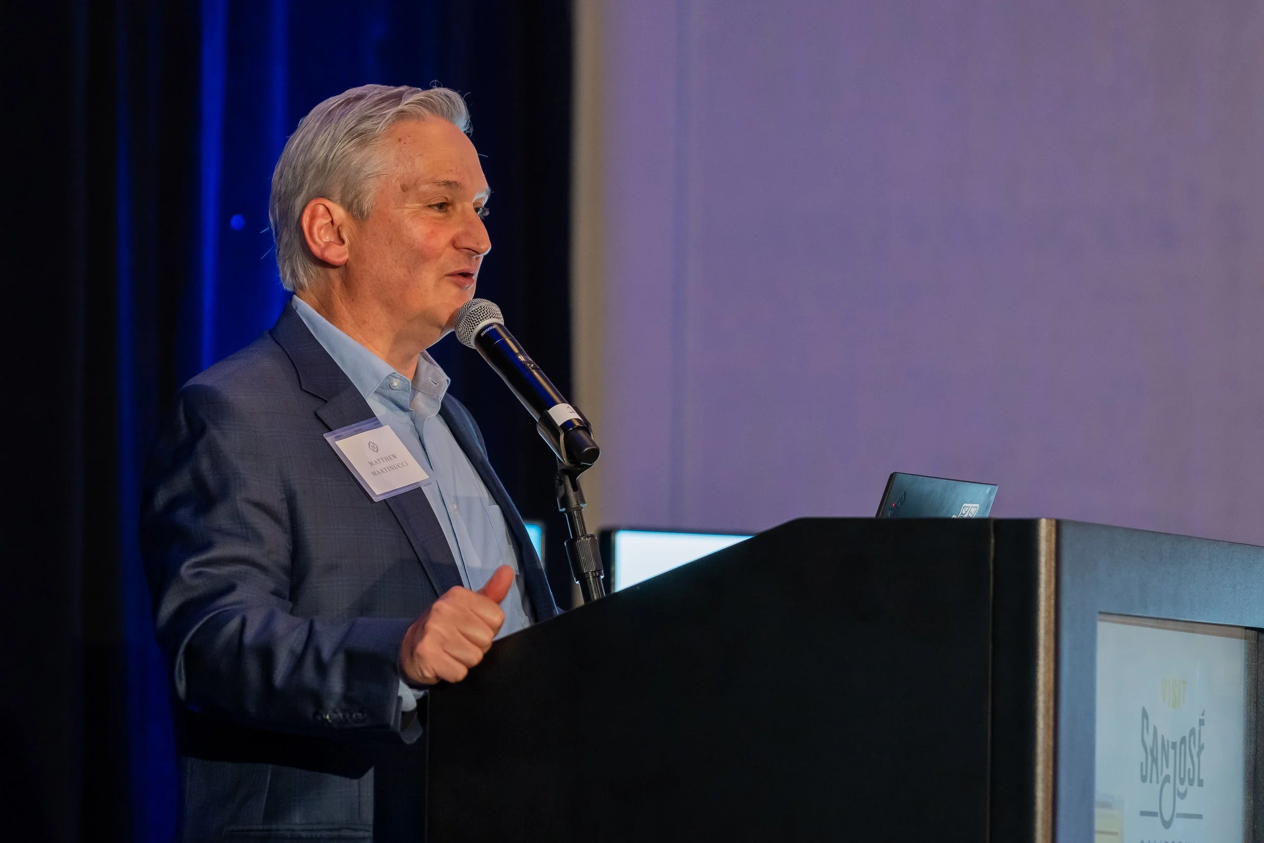 A man in a suit speaking into a microphone at a conference or presentation.