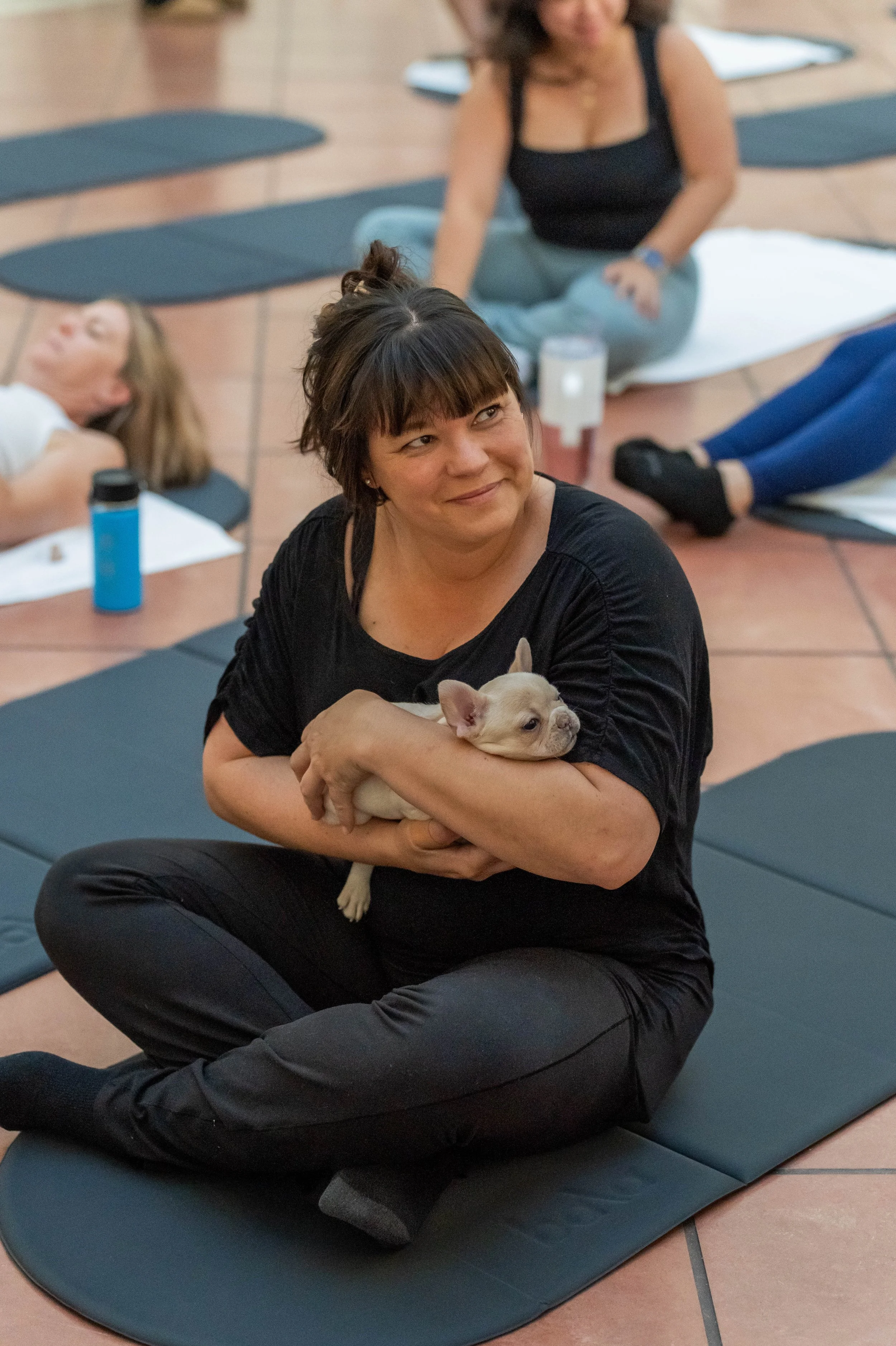 A woman with dark hair and bangs sitting cross-legged on a yoga mat, holding a small, light-colored French Bulldog puppy in her arms during a seated indoor yoga class. Several other participants are visible in the background, some lying on yoga mats,